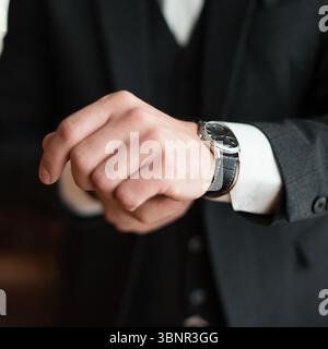 Nahaufnahme einer Hand eines Mannes, die eine Armbanduhr justiert. Die Uhr verfügt über ein silbernes Gehäuse und ein schwarzes Lederband. Er trägt einen dunklen Anzug. Stockfoto