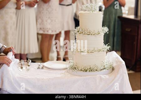 Eine makellose weiße Hochzeitstorte, drei Ebenen hoch, mit kleinen weißen Blumen verziert, sitzt auf einem Tisch. Gäste in weißen Kleidern sind im Hintergrund verschwimmen Stockfoto