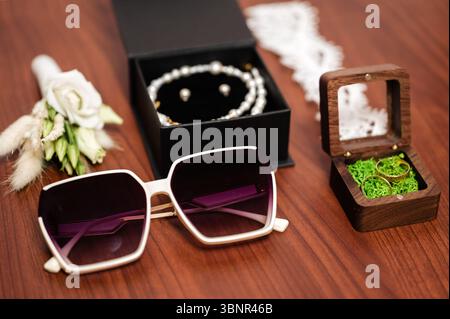 Ein stilvolles Arrangement von wichtigen Hochzeitsmaterialien: Elegante Sonnenbrillen, Perlenschmuck in einer Box und Eheringe in einem Holzetui. Die Szene ist eingestellt Stockfoto