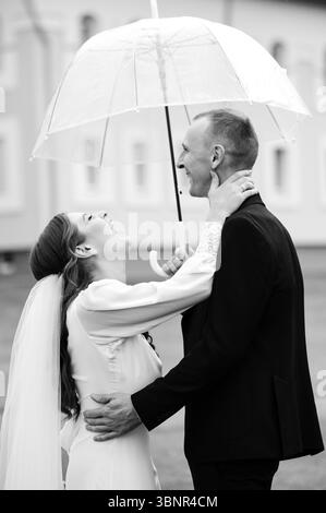 Schwarzweiß-Foto einer Braut und eines Bräutigams, die sich unter einem Regenschirm umarmen. Sie lacht, er lächelt. Eine romantische und intime Hochzeitsszene. Stockfoto