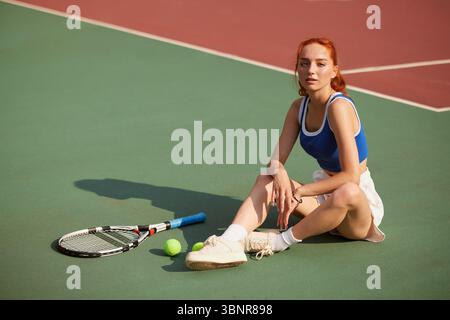 Sportliche junge Frau, die nach dem Training auf dem Tennisplatz sitzt und Schläger und Tennisbälle während des Sommertrainings hält Stockfoto
