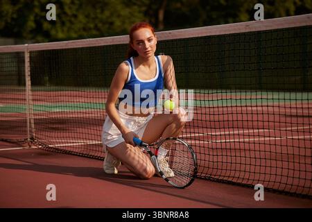 Junge Athletin kniet auf dem Tennisplatz mit Ball und Schläger, konzentriert während des Solo-Outdoor-Trainings Stockfoto