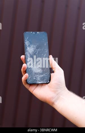 Eine vertikale Aufnahme einer Hand, die ein Mobiltelefon hält, mit einem stark gerissenen und gebrochenen Glasbildschirm, die eine beschädigte Kommunikation veranschaulicht. Stockfoto