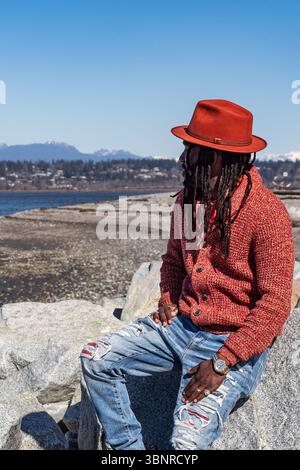 Schwarzer Mann mit Dreadlocks und Hut im Freien, genießt den Meerblick. Junger gutaussehender afro-amerikanischer Mann in stilvollem Outfit und hat Street Foto, Editorial V Stockfoto