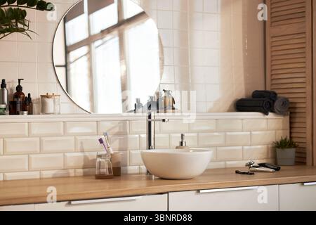 Moderne Arbeitsplatte im Badezimmer mit Hautpflegeprodukten, Seifenspender, elektrischem Rasierer, Zahnbürsten aus Glas, gerollten Handtüchern und Topfpflanzen, die Schönheit und Hautpflege widerspiegeln Stockfoto