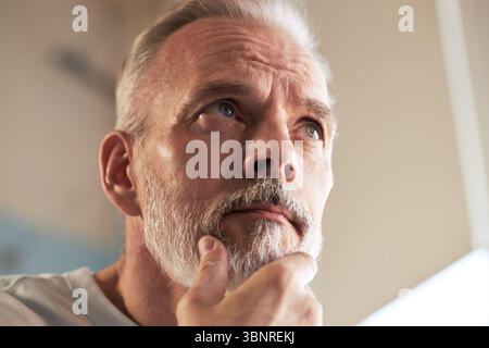 Porträt eines kaukasischen Mannes mittleren Alters, der den Bart berührt und nachdenklich nach oben blickt, zeigt das Hautpflegekonzept mit Nahaufnahme-Fokus auf Gesichtszüge und Pflege Stockfoto