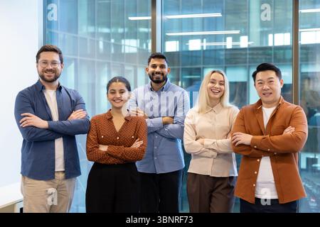 Eine Gruppe verschiedener Geschäftsleute lächelt und steht zusammen, mit überkreuzten Armen, in der Nähe eines Fensters. Stockfoto
