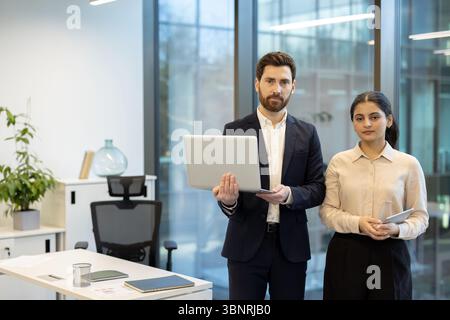 Ein Mann und eine Frau in einem Büro, der Mann in der Hand eines Laptops und die Frau ein Tablet, beide blicken nach vorne. Stockfoto