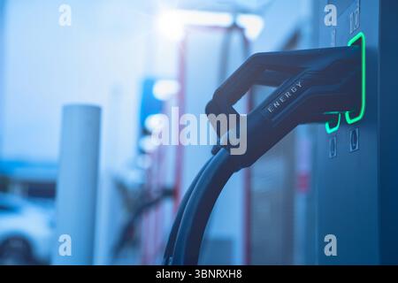 Nahaufnahme des EV-Ladeanschlusses, der an die Elektrofahrzeugstation angeschlossen ist, mit leuchtenden Lichtern in modernem Blauton für ein futuristisches Energiekonzept Stockfoto