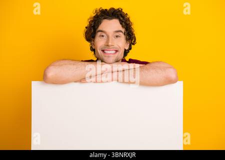 Fröhlicher junger Mann, der sich auf eine weiße Tafel vor einem hellgelben Hintergrund stützt und Glück und Positivität zeigt Stockfoto