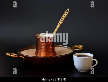 Ein Kupfertürke mit Kaffee steht in der Mitte eines Tabletts mit Sand, daneben befindet sich eine weiße Keramikschale mit einem Fertiggetränk auf schwarzem Hintergrund. Schließen- Stockfoto