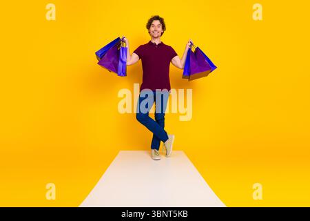 Fröhlicher junger Mann, der Einkaufstaschen auf einem bunten gelben Hintergrund hält, der Freude am Einkaufen und Stil zeigt Stockfoto