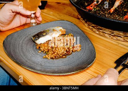 Ein Teller mit Reis und einem Stück Fisch wird auf einem Holztisch serviert, neben einer Hand mit einem Glas Bier und einer großen Paella-Pfanne mit Garnelen im B Stockfoto