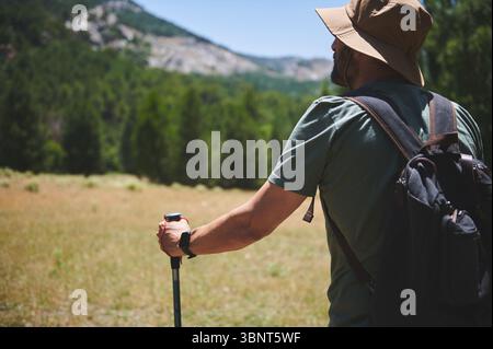 Mann ausgestattet mit Rucksack und Hut Wandern durch malerisches bergiges Gelände umgeben von üppigem Grün im Sommer, symbolisiert Abenteuer und Erkundungen Stockfoto