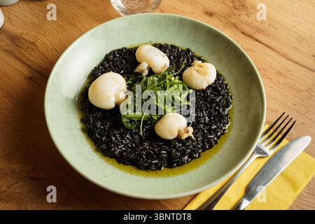 Ein Gourmet-Teller mit Risotto aus schwarzer Tintenfisch-Tinte, garniert mit gebratenem Tintenfisch und einer frischen Rucola-Garnitur. Der leuchtende Grün- und Tiefschwarz-Kontrast b Stockfoto
