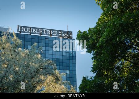 Lodz, Polen – 2. Juli 2025: Schild mit dem Doubletree by Hilton Hotel auf einem modernen Glasgebäude, teilweise von Bäumen verdeckt und von warmem Abendlicht beleuchtet. Stockfoto