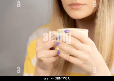 Eine Frau in einem gelb-weißen T-Shirt trinkt Kaffee mit Milch aus einer blassgelben Tasse. Der untere Teil ihres Gesichts ist in einer extremen Nahaufnahme zu sehen. Stockfoto