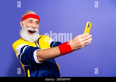 Fröhlicher Seniorenmann, der Selfie macht, während er Retro-Sportswear und Stirnband trägt, der Nostalgie und Glück ausstrahlt Stockfoto