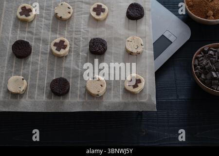 Vorderansicht des Keksteigs auf Backpapier mit Schüssel mit Schokoladenchips, brauner Zuckerschale und digitaler Skala auf schwarzer Holzoberfläche Stockfoto