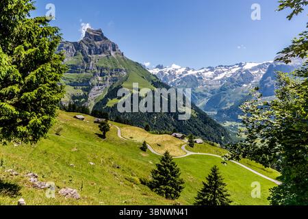 Hahnen (2.606 m ü. M.) gilt als Hausberg der Stadt Engelberg. Er liegt in den Urner Alpen im Kanton Obwalden und erhebt sich deutlich über dem Engelbergtal. Eine idyllische Landschaft in oberer Flühmatt, Engelberg, Obwalden, Schweiz. Das Bild zeigt grüne Almwiesen mit traditionellen Holzhütten und einer gewundenen Straße, umgeben von majestätischen Bergen, von denen einige noch mit Schnee bedeckt sind. Es ist eine malerische Szene, die die Schönheit der Schweizer Alpen im Sommer fängt Stockfoto