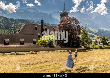 Sommer in Engelberg: Frauen wenden Heu vor den Mauern des Benediktinerklosters in Engelberg. Zwei Personen hacken Heu auf einem Feld vor dem Benediktinerkloster Engelberg in Engelberg, Obwalden, Schweiz. Sie arbeiten an der traditionellen Heuernte, während ein Gleitschirmflieger über die majestätischen Berge im Hintergrund schwingt Stockfoto