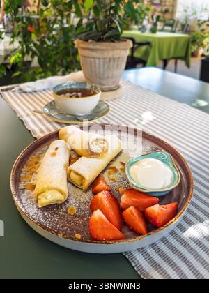 Ein köstliches süßes Frühstück mit Käsecrepes (Blini), frischen Erdbeeren und Sauerrahm, serviert mit Tee in einem gemütlichen Café.Ein gesundes und leckeres Stockfoto
