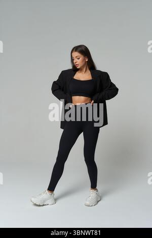 Selbstbewusste Frau in dunkler Sportkleidung und Turnschuhen posiert mit Händen auf der Hüfte. Sie trägt eine schwarze Jacke über einem passenden Crop-Top und Leggings. Stockfoto