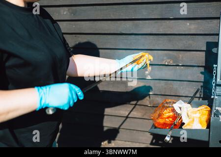 Eine Person verwendet eine Zange, um Speisen auf einem Grill zuzubereiten und draußen zu kochen. Eine Frau mit blauen Handschuhen hält rohes Fleisch mit einer Zange in der Nähe eines Grills. Stockfoto