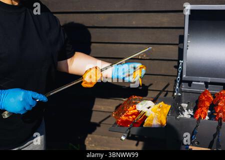 Eine Person verwendet eine Zange, um Speisen auf einem Grill zuzubereiten und draußen zu kochen. Eine Frau mit blauen Handschuhen hält rohes Fleisch mit einer Zange in der Nähe eines Grills. Stockfoto