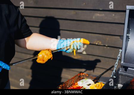 Eine Person verwendet eine Zange, um Speisen auf einem Grill zuzubereiten und draußen zu kochen. Eine Frau mit blauen Handschuhen hält rohes Fleisch mit einer Zange in der Nähe eines Grills. Stockfoto