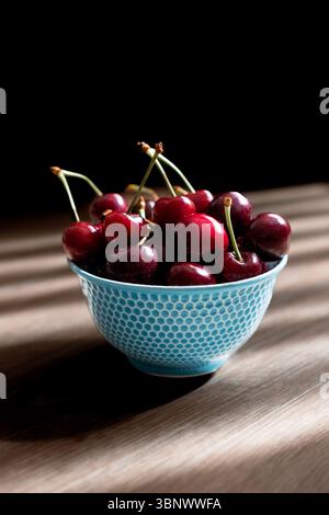 Schüssel mit Reifen roten Kirschen in einer blau strukturierten Schüssel, beleuchtet von natürlichem Licht mit starken Schatten auf einem Holztisch und dunklem Hintergrund Stockfoto