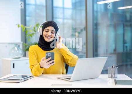 Eine Frau, die einen Hijab trägt, lächelt, während sie in einem Büro mit Kopfhörern auf ihr Telefon schaut. Stockfoto