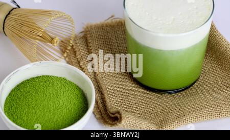 Cremiger Matcha-Latte mit Bambus-Schneebesen und leuchtendem grünem Matcha-Pulver auf rustikalem Sackleinen, minimalistischer Overhead-Stil Stockfoto