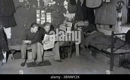 1946, historische, ausländische Arbeiter in ihrem Bett im Volkswagen-Werk Wolfsburg, zwei sitzen auf einem Lagerbett und lesen eine Zeitung, möglicherweise junge lettische Männer. Sozialkekse von McVitie & Price Ltd. Und Dosen von Alaska-Lachs unter dem Bett. Unter der Kontrolle der britischen Armee begann ab Juni 1945 die Produktion von Volkswagen-Autos und viele Flüchtlinge und Migranten kamen aus den ehemaligen deutschen Gebieten in Osteuropa nach Wolfsburg, um Nahrung und Wohnung zu suchen. Für die meisten Werksarbeiter waren die Lebensbedingungen grundlegend. Stockfoto