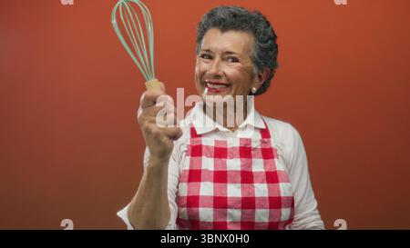 Eine ältere Frau mit grauem Haar in einer roten karierten Schürze hält fröhlich einen Schneebesen vor einem orangefarbenen Hintergrund und zeigt ihre Leidenschaft für Kochen und Küche Stockfoto
