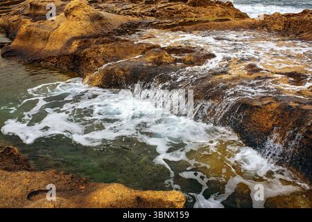 Meerwasser fließt sanft über natürliche Küstenfelsen und bildet eine flache Kaskade mit weißem Schaum. Malerische und dynamische Meereslandschaft Stockfoto