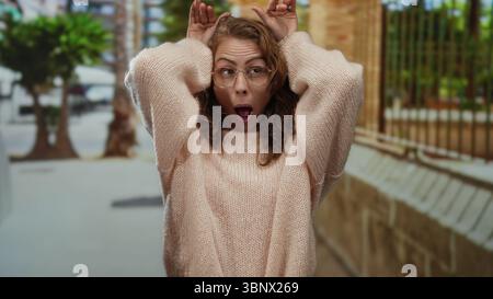 Frau macht lustige Gesichter in einer urbanen Straßenumgebung, die mit Brille und lockigem Haar im Freien in einer städtischen Umgebung mit Autom. Verspielt ausdrückt Stockfoto