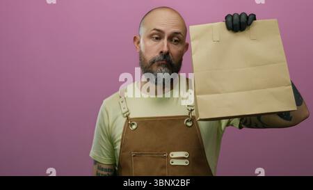 Kahler bärtiger Mann in Schürze, der Papiertüte mit ausdruckslosem Gesicht vor rosa Hintergrund hält. Stockfoto