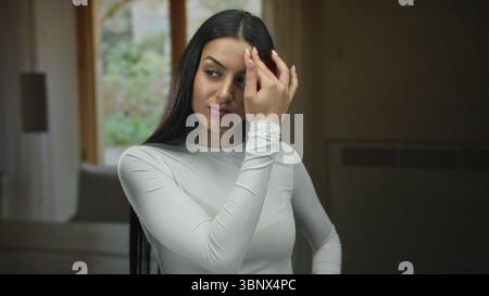 Frau in weiß drinnen, die nachdenklich oder besinnlich wirkt, mit Hand auf der Stirn vor einem weichen Hintergrund, was einen introspektiven Moment in sich andeutet Stockfoto