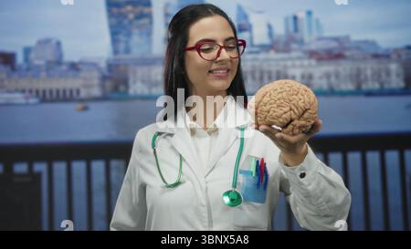 Frau Ärztin mit Hirnmodell an der Strandpromenade mit Stethoskop in Uniform im Freien, selbstbewusst lächelnd. Stockfoto