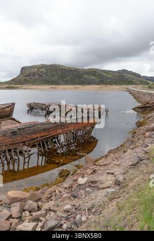 Verrostete Schiffswracks aus Holz und Metall tauchten teilweise in flachen Küstengewässern vor einem Hintergrund von felsigen Hügeln und bewölktem Himmel Stockfoto