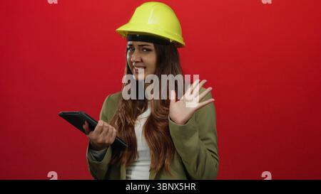 Die hispanische Ingenieurin mit gelbem Schutzhelm hält eine Tablette vor einem kräftigen roten Hintergrund und drückt Frustration, Energie und Entschlossenheit aus Stockfoto