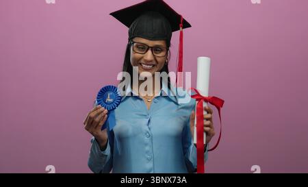 Lächelnde Frau in Abschlusskleidung mit Diplom und Band vor rosa Hintergrund feiert akademische Leistungen. Stockfoto