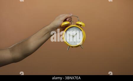 Die Hand des kaukasischen Mannes hält den gelben Wecker an der braunen Wand, zeigt, isoliert, zeigt Zeitdetails mit Fokus auf die Morgeneinstellung. Stockfoto