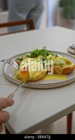 Person genießt köstliche Eier benedict mit Avocado drinnen in einem gemütlichen Restaurant mit einer Gabel und einem Messer auf einem weißen Tisch mit frischen Zutaten A Stockfoto