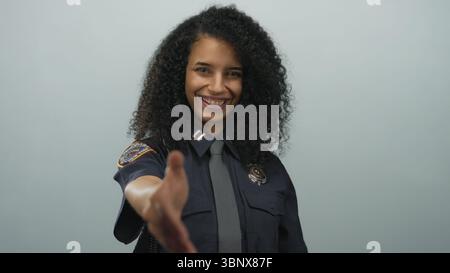 Hispanische Polizistin in Uniform streckt einen Handschlag und lächelt selbstbewusst vor weißem Hintergrund. Stockfoto