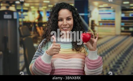 Die lächelnde Frau hält das Herz, während sie im Fitnessstudio mit einem farbenfrohen Pullover die Daumen nach oben drückt und im Fitnesscenter positiver wirkt. Stockfoto