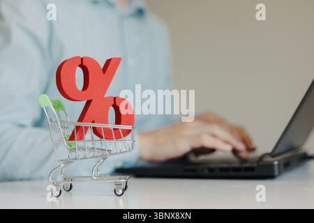 Rotes Prozentzeichen im kleinen Einkaufswagen auf dem Tisch. Mann, der im Hintergrund einen Laptop benutzt. Verkauf, Black Friday, Online-Shopping-Konzept. Stockfoto