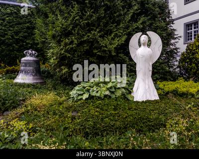 Eine historische Kirchenglocke und eine moderne Engelsstatue aus weißem Marmor stehen im Garten der Abtei Engelberg, gegründet 1120 von Konrad von Sellenbüren und eine der ältesten Benediktinerabteien der Schweiz. Am Aeschiweg in Engelberg, Kanton Obwalden, Schweiz, verbindet die Szene klösterliche Tradition mit zeitgenössischer religiöser Kunst. Stockfoto