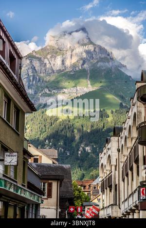 Blick von Engelbergs Altstadt auf die 2.606 Meter hohen Hahnen. Der Gipfel befindet sich etwa vier Kilometer östlich des Ortskerns und ist über anspruchsvolle Alpenrouten zu erreichen. Engelberg, Schweiz: Ein beeindruckender Blick auf die majestätischen Schweizer Alpen von der Bahnhofstraße. Der Berggipfel ist teilweise von Wolken umhüllt, während traditionelle Gebäude die Szene umrahmen. Schweizer Flaggen zieren die Straße und betonen die lokale Identität und die alpine Umgebung Stockfoto